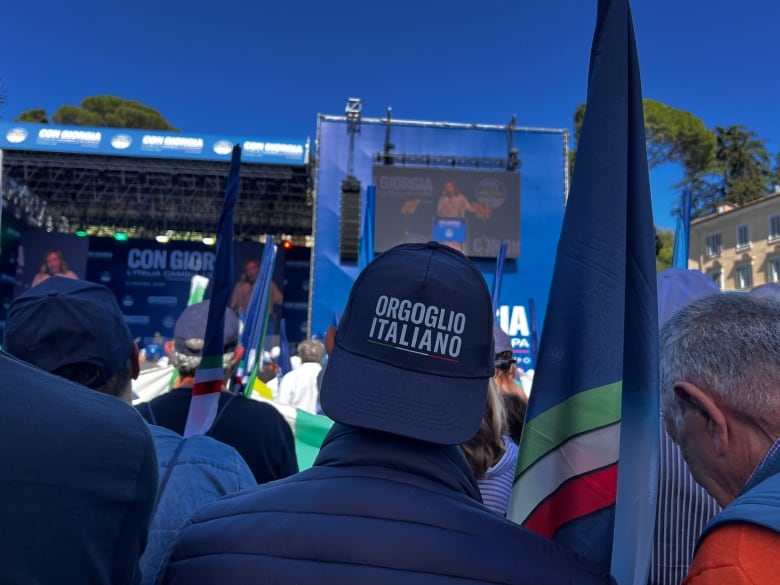 People in a crowd look toward a screen that shows an image of a person speaking.