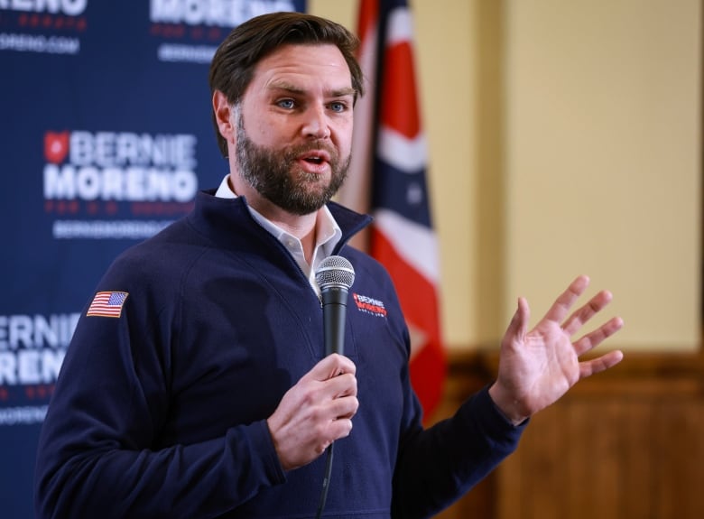 A bearded man wearing a collared fleece pullover holds a microphone and speaks indoors.