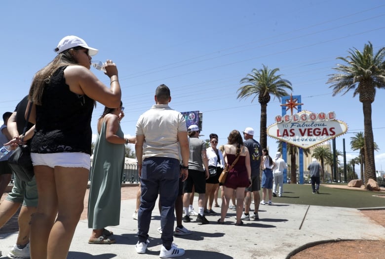 A woman wearing a white ball cap, a dark top and white shorts drinks from a bottle as she stands in a line of people beside a sign that reads "Welcome to Fabulous Las Vegas Nevada."